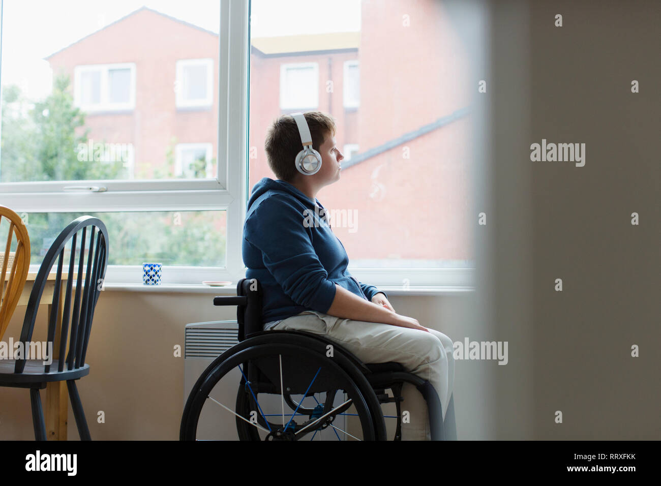 Nachdenkliche junge Frau im Rollstuhl Musikhören mit Kopfhörern mit Fenster Stockfoto