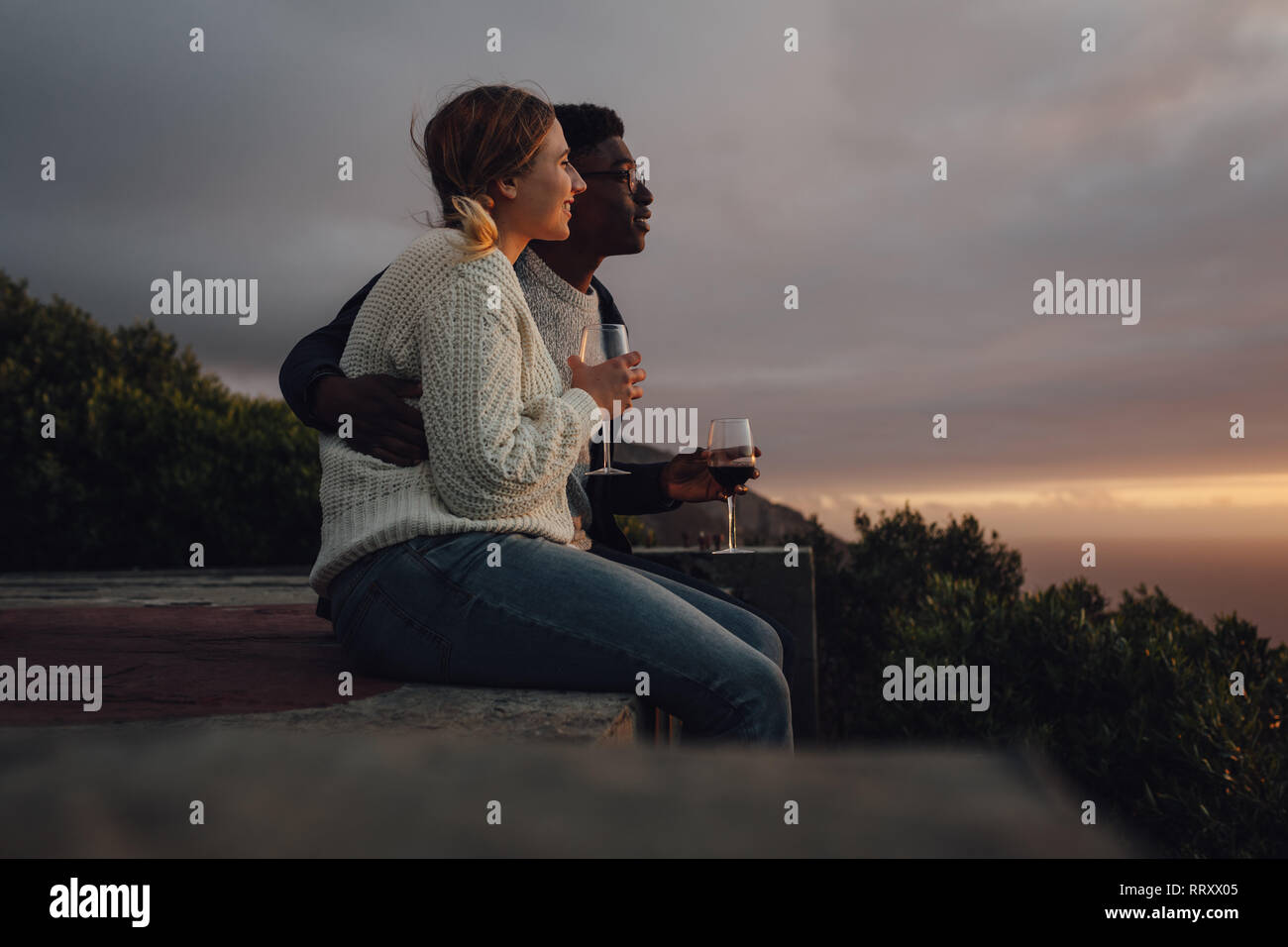 Seitenansicht der romantischen interracial Paar bewundern Sie den Sonnenuntergang. Der Mann und die Frau sitzen zusammen mit Weingläsern. Stockfoto