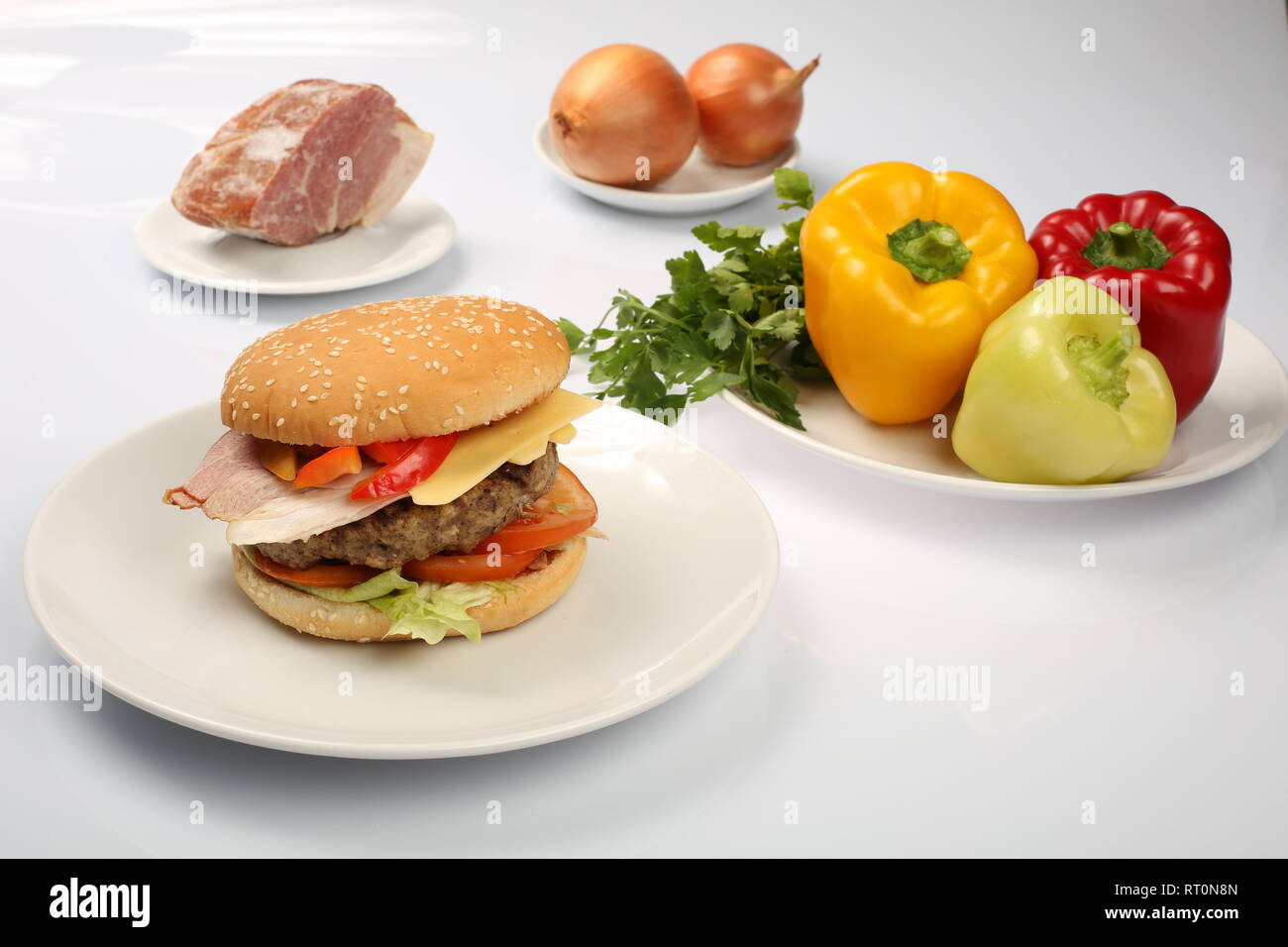 Cheeseburger mit Speck und Rindfleisch Patty auf einem Teller mit Gemüse, Paprika, Tomaten, Zwiebeln, Gurken, Petersilie und Salat auf einem weißen Hintergrund, s Stockfoto