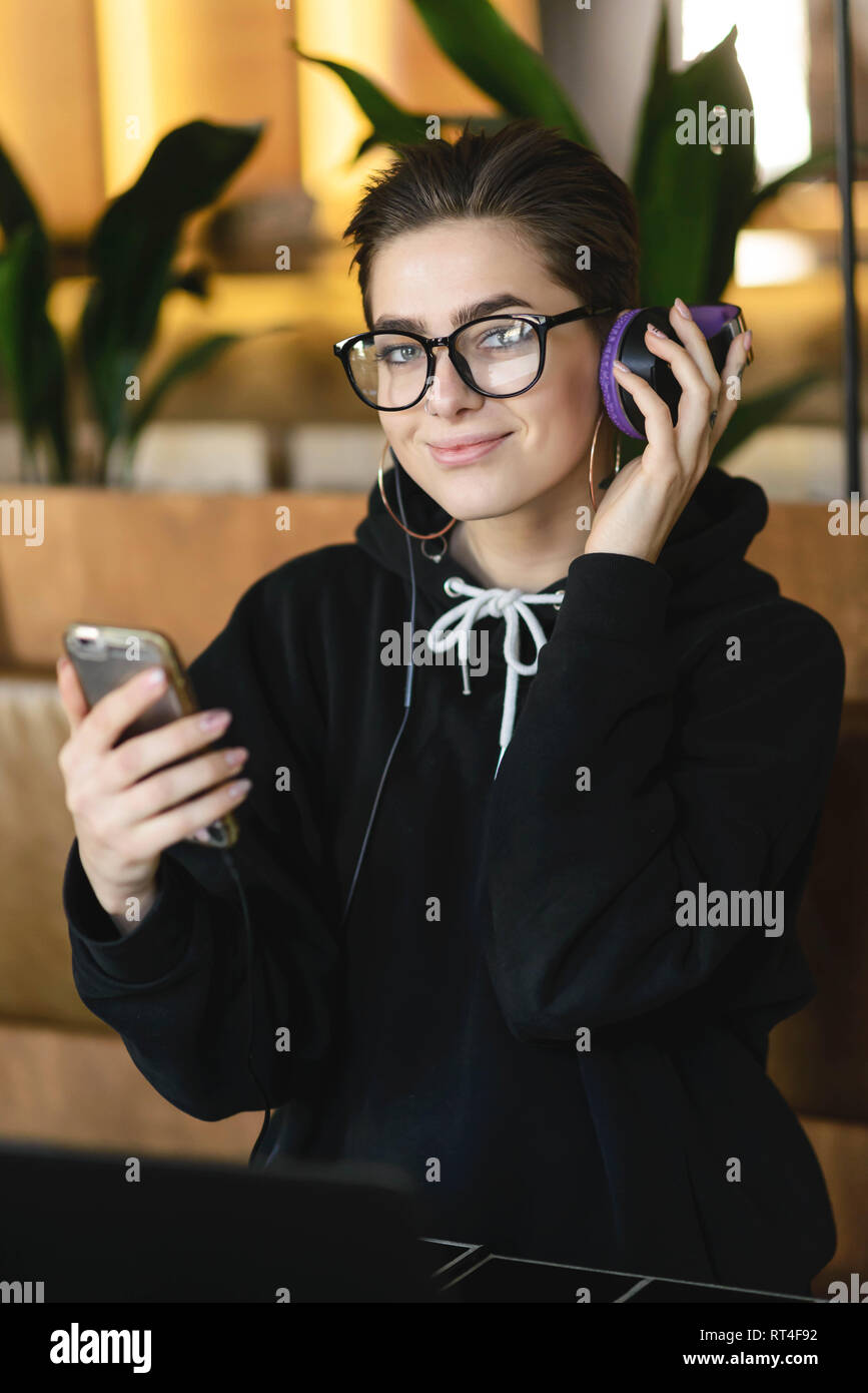 Portrait von Kurzhaar hipster Girl in Brillen tragen Black Hoody hören Musik auf Ihr Smartphone, während im Cafe sitzen Stockfoto