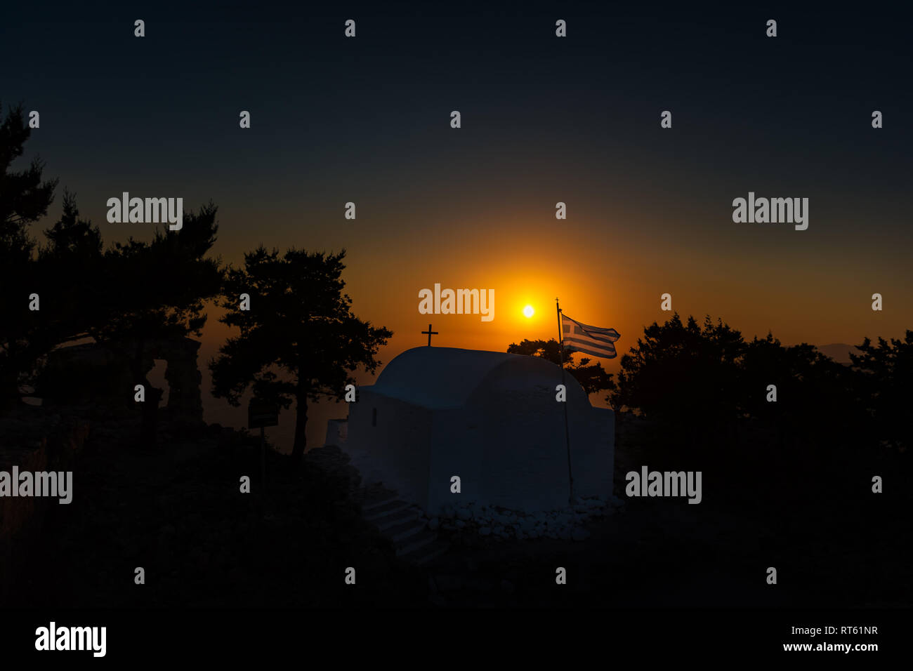 Blick auf griechische Flagge und die orthodoxe Kirche erbaut auf den Ruinen der Burg von Monolithos bei Sonnenuntergang (Rhodos, Griechenland) Stockfoto