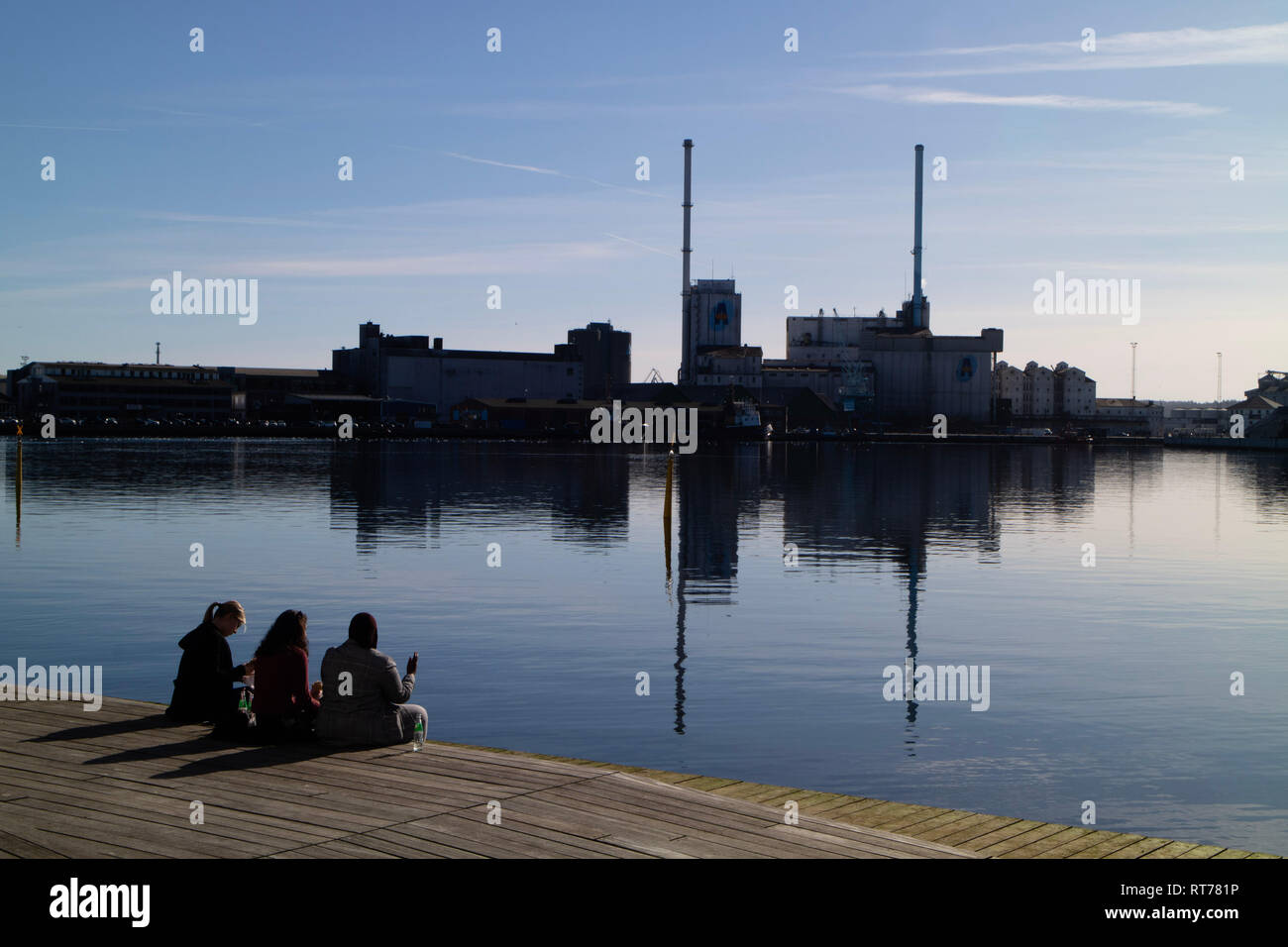 Studenten entspannende außerhalb der Universität Aarhus Navitas Gebäude neben Hafen während einer Periode der ungewöhnlich warmen Witterung im Februar 2019. Die Temperaturen erreichen 16 C obwohl die Februar durchschnittlich 6 ist C. Eine Landwirtschaft der industriellen Fabrik ist im Hintergrund. Aarhus, Dänemark. Februar 2019. Stockfoto