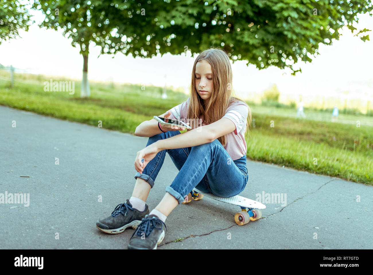 Jugendlich Mädchen 11-14 Jahre alt und sitzt in Händen der Skate. Im Sommer Stadt in lässigen Jeans und pink T-Shirt. Kommunikation im Internet, in den Händen smartphone Stockfoto