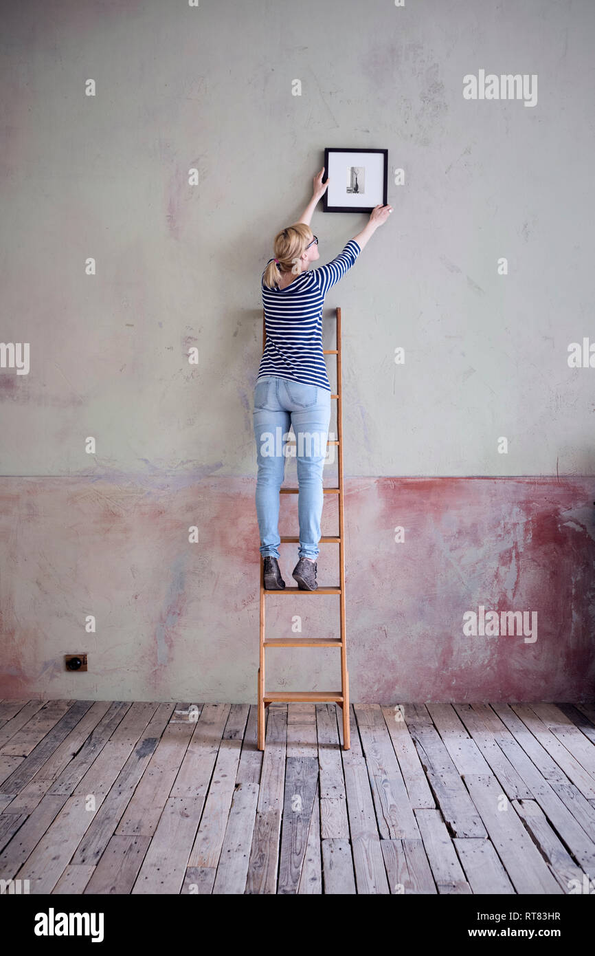 Rückansicht der Frau auf Leiter aufhängen Bilderrahmen in einem unrenovierten Zimmer eines Loft Stockfoto