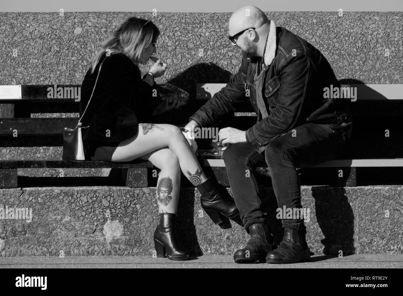 Schwarzweiß-Bild einer jungen Frau mit tätowierten Beinen auf einer Bank sitzen Essen mit einem männlichen Begleiter. Stockfoto