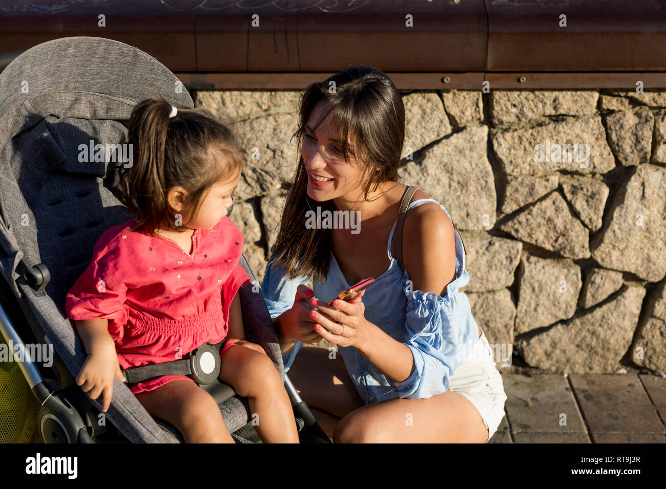 Die Mutter kniet neben Tochter im Kinderwagen sitzen, am Smartphone suchen Stockfoto