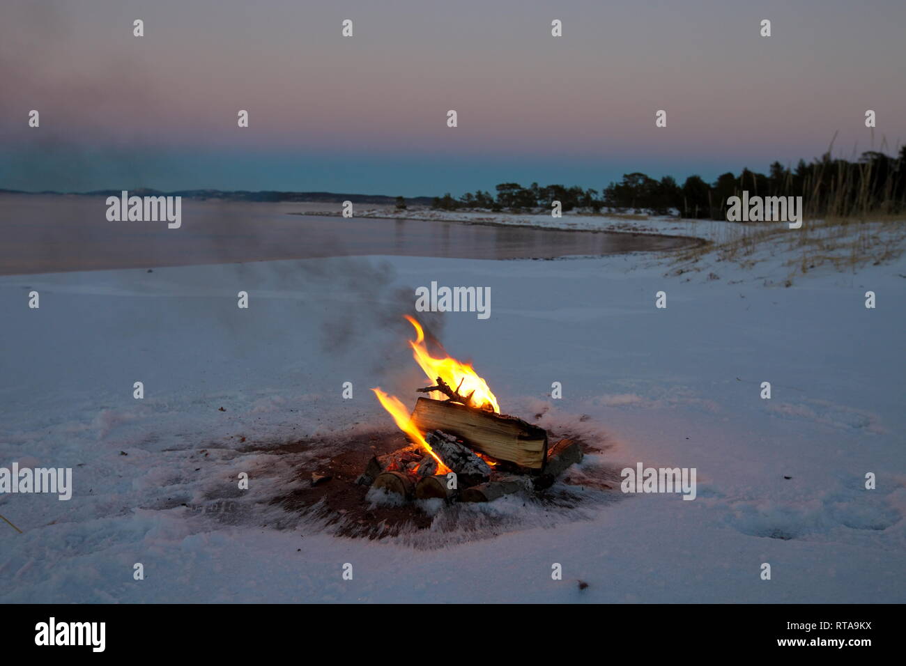 Ein Lagerfeuer brennt am Ufer einer Bucht der Ostsee. Es ist Sonnenuntergang an einem Wintertag, und der Strand ist mit Schnee bedeckt. Stockfoto