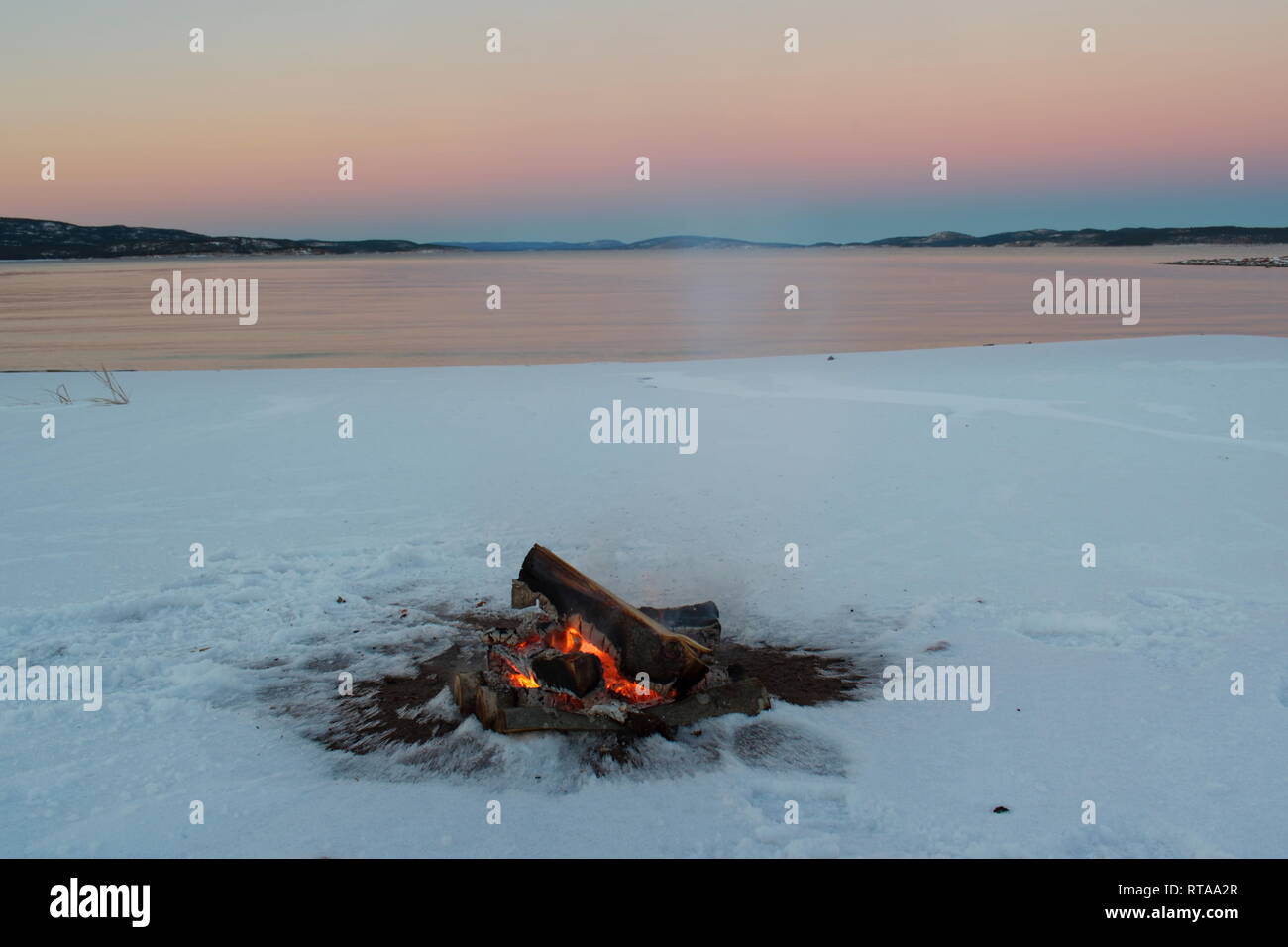 Ein Lagerfeuer brennt am Ufer einer Bucht der Ostsee. Es ist Sonnenuntergang an einem Wintertag, und der Strand ist mit Schnee bedeckt. Stockfoto