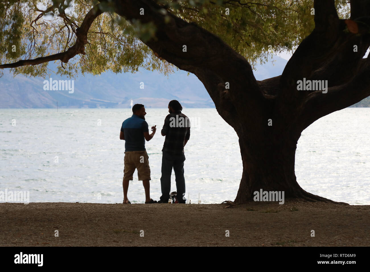 Zwei Männer sprechen und Trinken auf dem Kiesstrand bei Queenstown, Lake Wakatipu, Neuseeland Südinsel Stockfoto