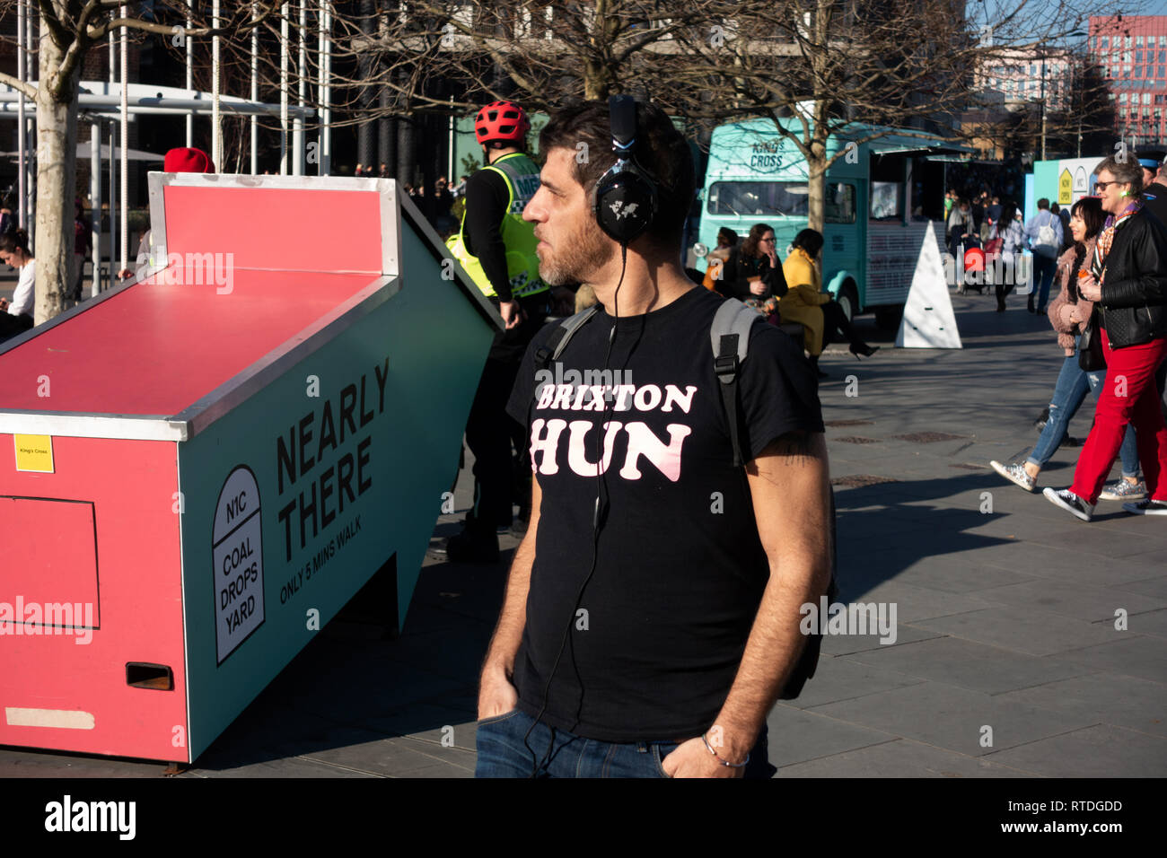 Junger Mann Kopfhörer tragen in ein Schwarzes T-Shirt mit Slogan Brixton Hun zu Fuß neben 'Fast' 3D-Schild in der Nähe von Kings Cross, London Stockfoto