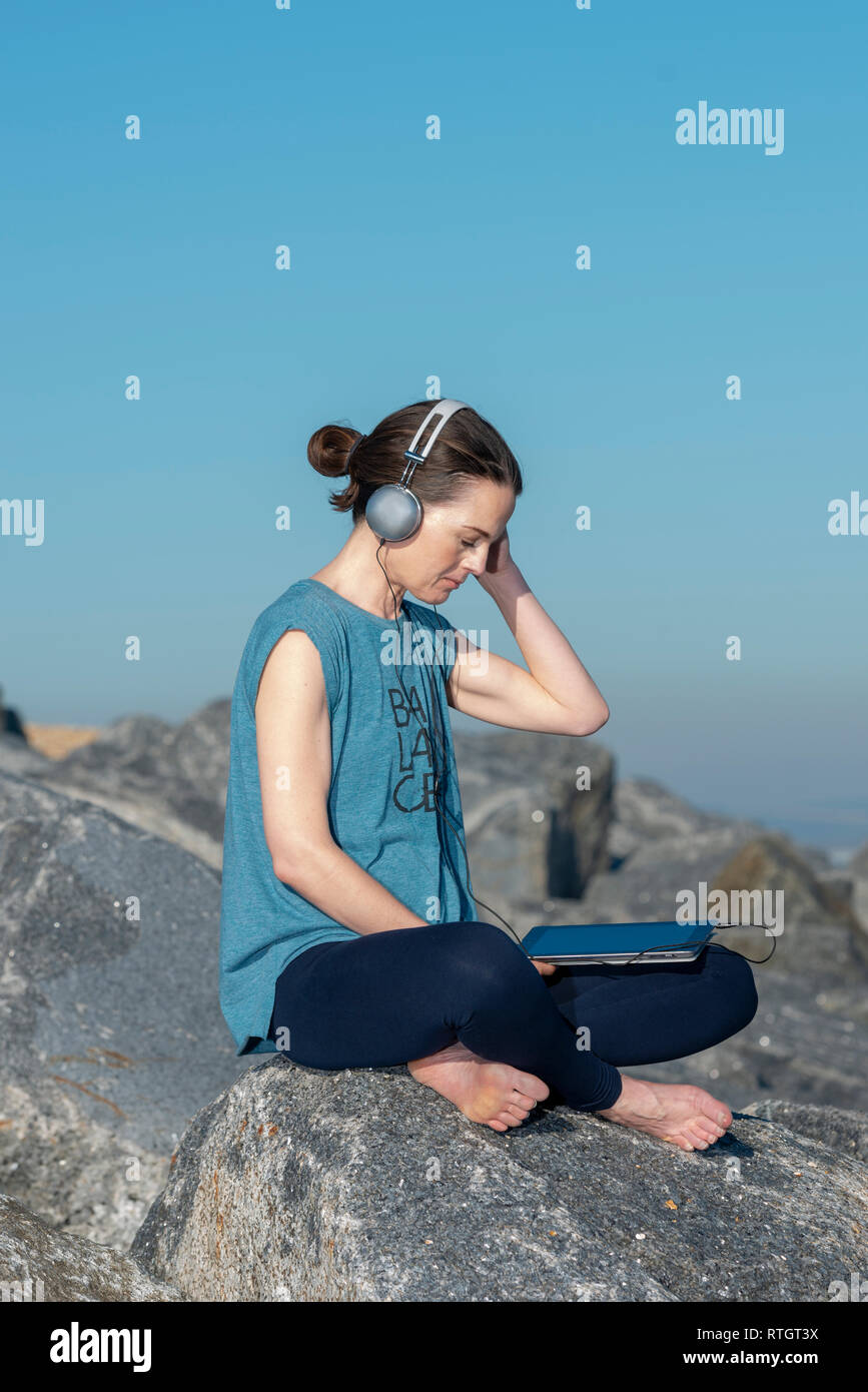 Frau sitzen auf den Felsen am Meer Musik auf Ihrem Tablet mit Kopfhörern Stockfoto
