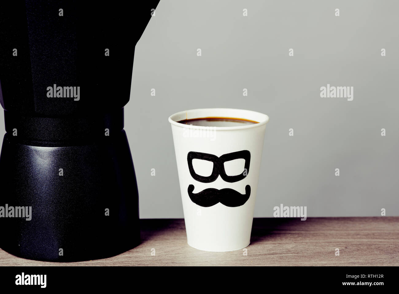 Nahaufnahme der Kaffeemaschine und eine Tasse mit Kaffee, mit ein paar Gläser und einen Schnurrbart in es gezeichnet, mit der Darstellung eines Menschen Gesicht, auf einem Holztisch Stockfoto