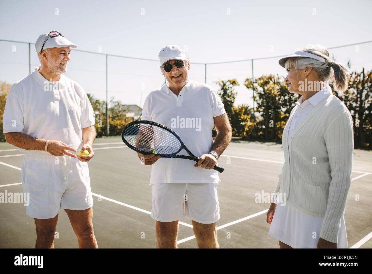 Freundliche Männer im Tennis tragen im Gespräch mit einer älteren Frau, die auf einem Tennisplatz. Lächelnd alten Mann, der mit seinen Spielkameraden Holding einen Tennisschläger. Stockfoto