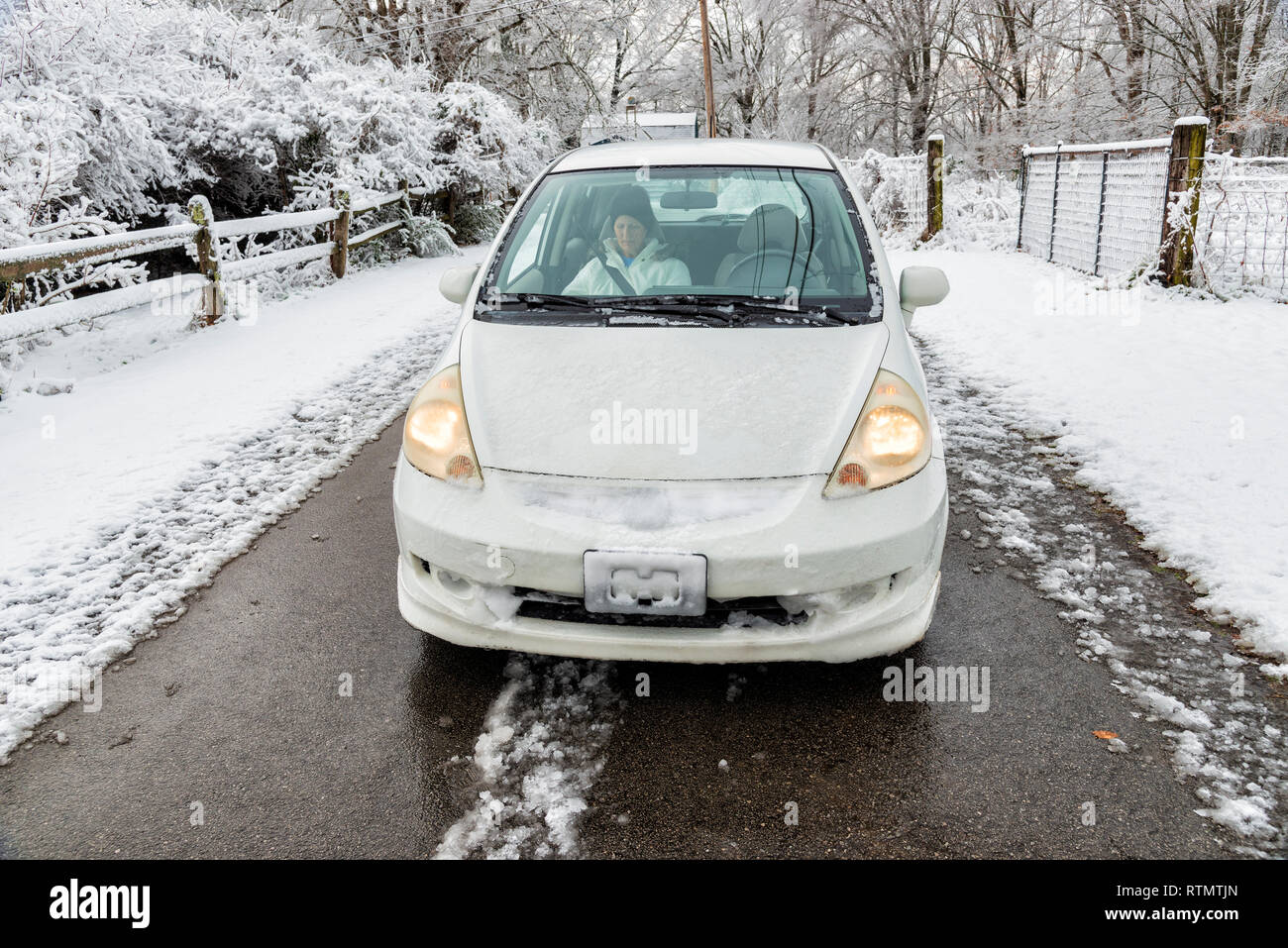 Horizontale geschossen von einem weißen Auto auf einer verschneiten Straße mit einer mittleren Alters Lady auf dem Beifahrersitz. Die Scheinwerfer eingeschaltet sind. Schnee ist schwerer zu beiden Seiten des Stockfoto