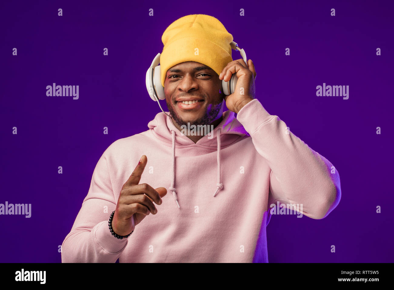 Junge afrikanische Mann in Hoodie, in der Sinn für Mode eines street Urchin, drehte sich ein wenig in der Kopfhörer auf der violette Hintergrund isoliert, holding Ohrhörer Stockfoto