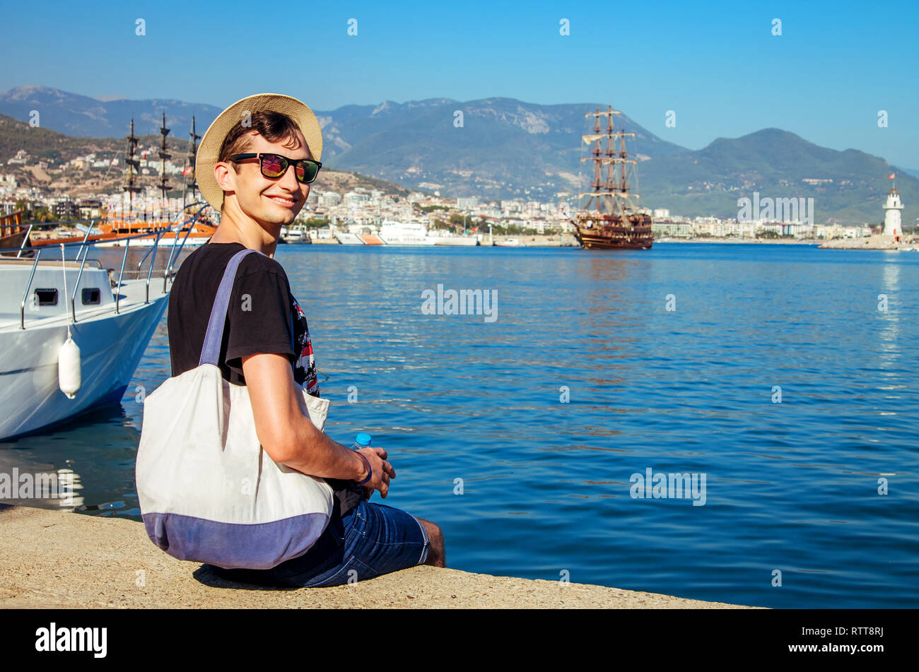 Junge glücklich Mann sieht die Alanya Hafen von Bergen umgeben ist. Lächelnd Reisende sitzend durch das Meer. Ferienhäuser Konzept Stockfoto Junge glücklich Mann sieht die Alanya Hafen von Bergen umgeben ist. Lächelnd Reisende sitzend durch das Meer. Ferienhäuser Konzept Stockfoto