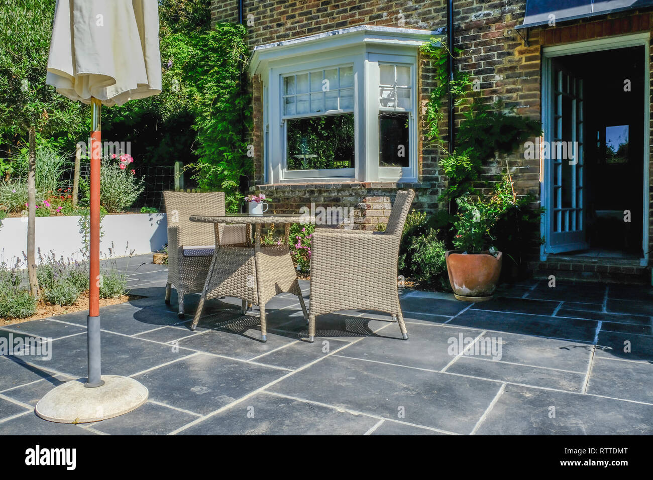 Korbstühle und Rohrgeflecht Tisch in der Mitte eines Schiefer Patio mit Pflanzen und Bäumen in Beeten und Töpfen. An einem sonnigen Sommertag mit niemand in der übernommen Stockfoto