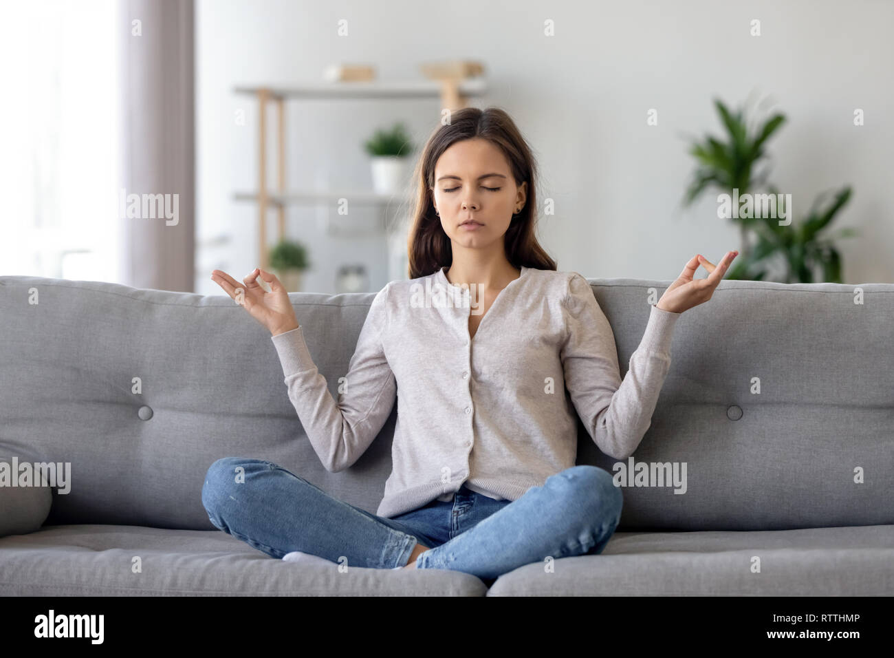 Ruhige junge Frau sitzt im Lotussitz auf dem Sofa, Meditieren zu Hause Stockfoto