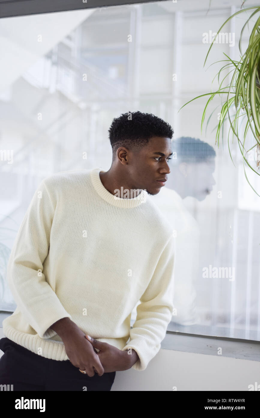 Porträt von einem gutaussehenden jungen männlichen Schwarzen stehen allein in einem Zimmer mit Fenstern und üppigen Pflanzen. Es ist ein Tower Block außerhalb Stockfoto