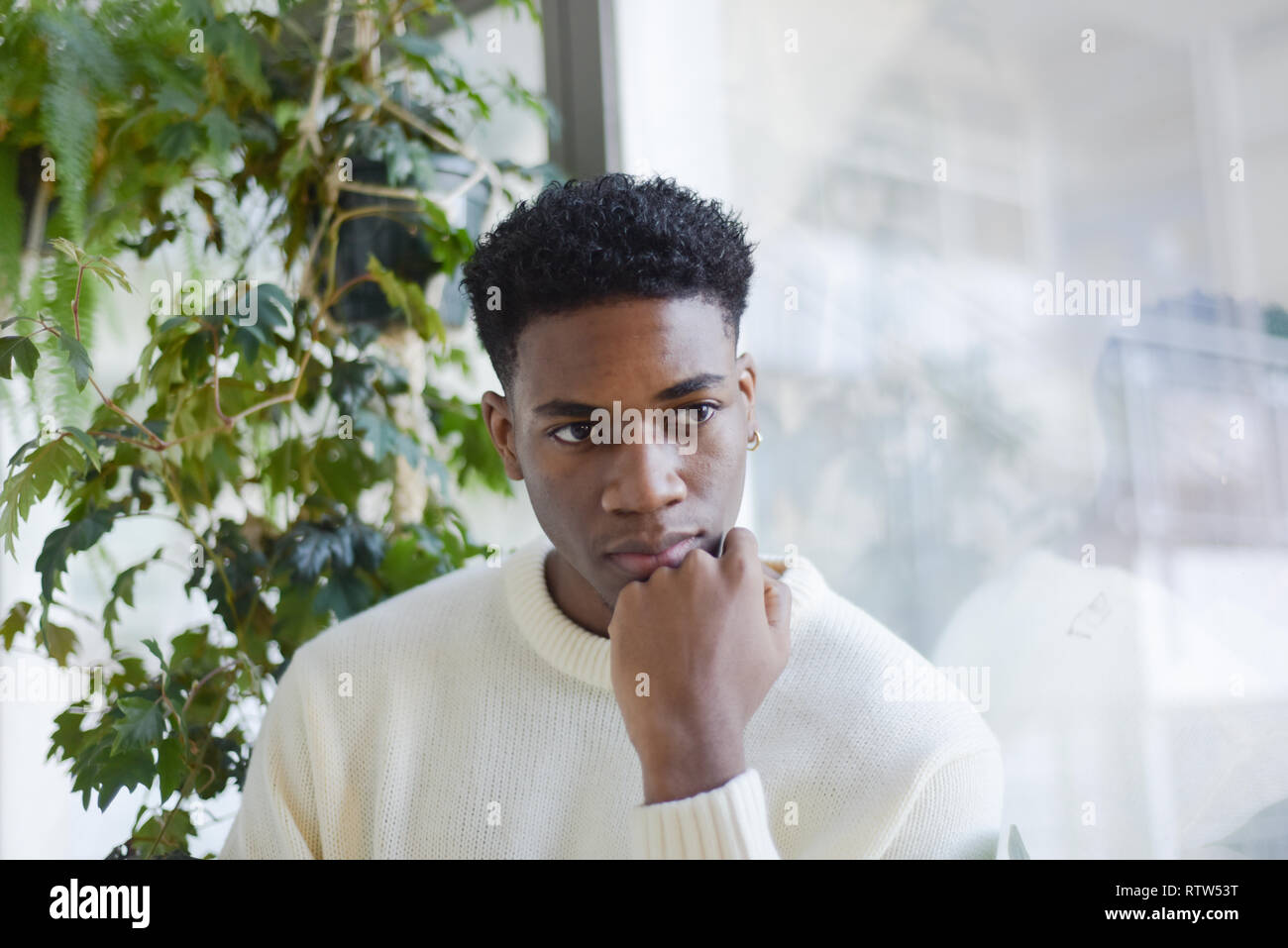 Porträt von einem gutaussehenden jungen männlichen Schwarzen stehen allein in einem Zimmer mit Fenstern und üppigen Pflanzen. Es ist ein Tower Block außerhalb Stockfoto