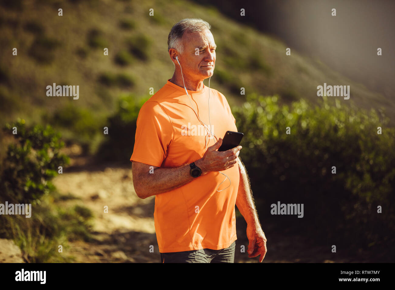 Älterer Mann in fitness Verschleiß hören die Musik auf Handy bei einem Spaziergang. Porträt eines Senior Fitness Mann auf einem hügeligen trail Track hören Stockfoto