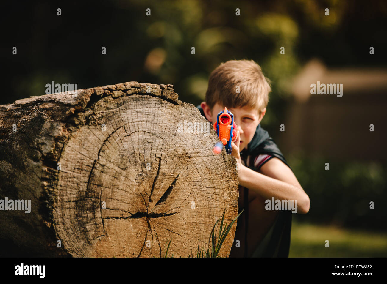 Lustige kaukasischen Kid in Kap Versteckt hinter einer mit spielzeugpistole in der Hand anmelden. Junge spielt mit spielzeugpistole im Freien. Stockfoto