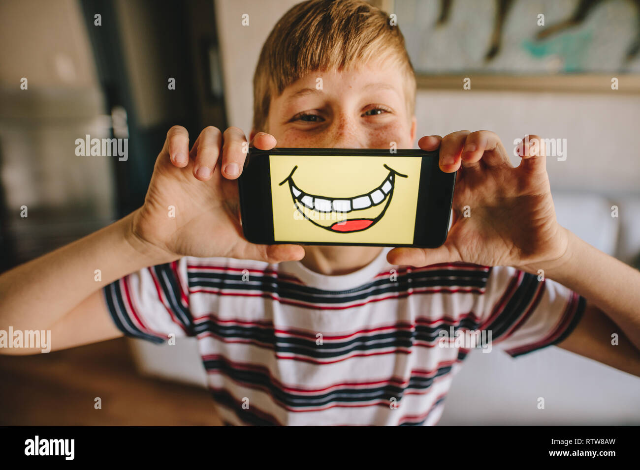 Junge Holding ein Smartphone vor seinem Gesicht mit smiley Bild auf dem Display. Junge vorgibt, zu Hause, glücklich zu sein. Stockfoto