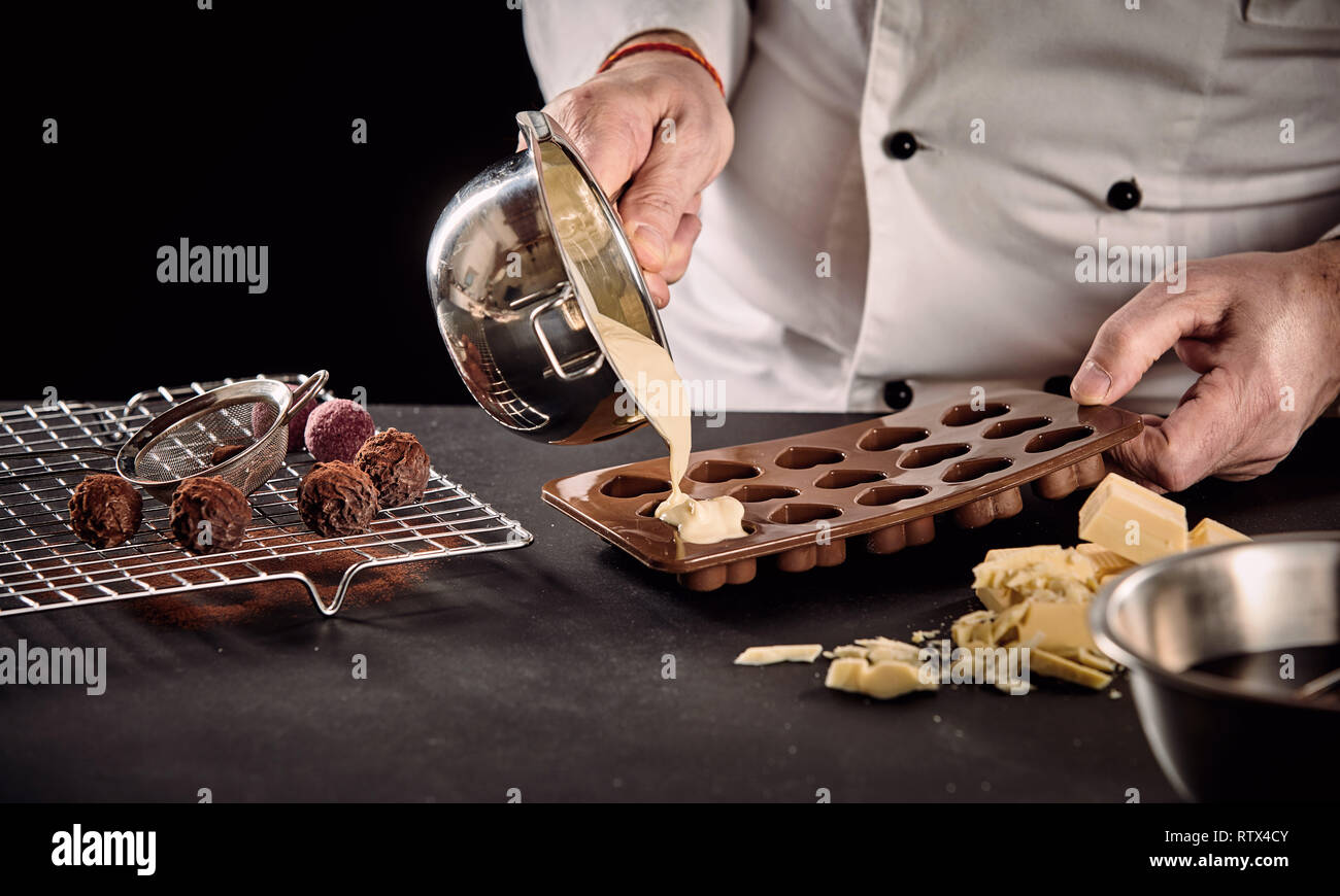 Koch oder Chocolatier gießen geschmolzene weiße Schokolade in Silikon Backformen aus einem Topf in einer Nahaufnahme auf seinen Händen Stockfoto
