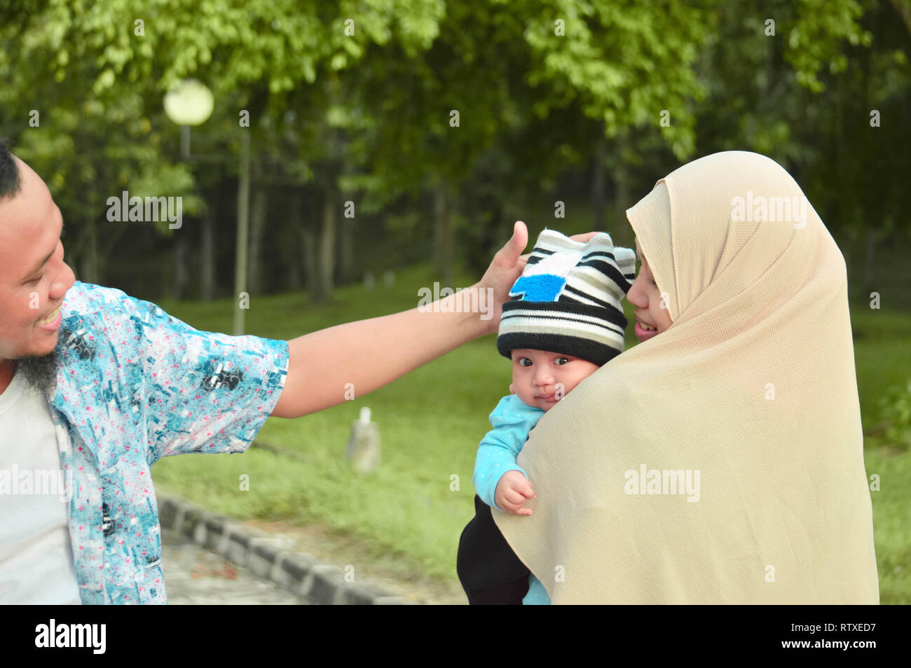 Asiatische muslimischen hijabi Mutter und Vater zu Fuß durch den Park mit Sohn im Kinderwagen, während seine Mutter kümmert sich um Ihr Baby Stockfoto