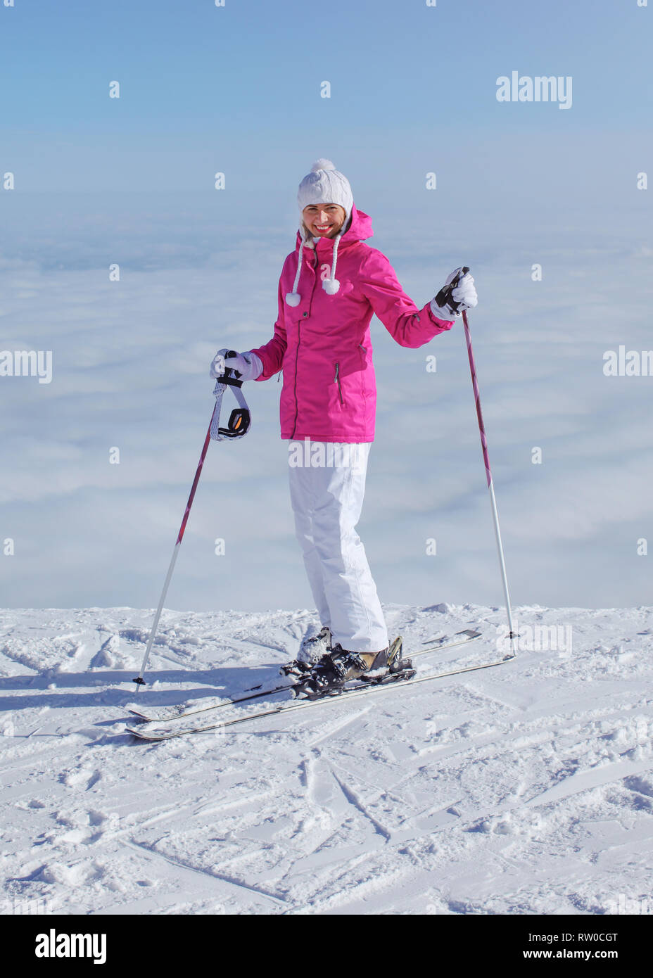 Frau in rosa Jacke, Ski Stöcke und Skier, am Rand des Hügels stehend, nur Wolken unter, lächelnd Stockfoto