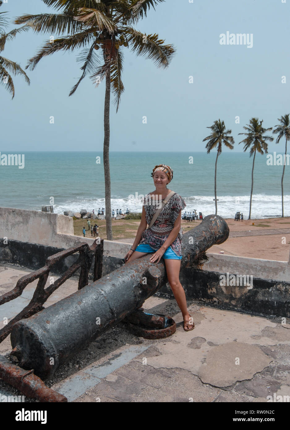 Ein Foto von einem Europäischen Frau auf eine riesige Kanone auf Elmina Castle posieren. Ozean können auf dem Hintergrund gesehen werden. Stockfoto