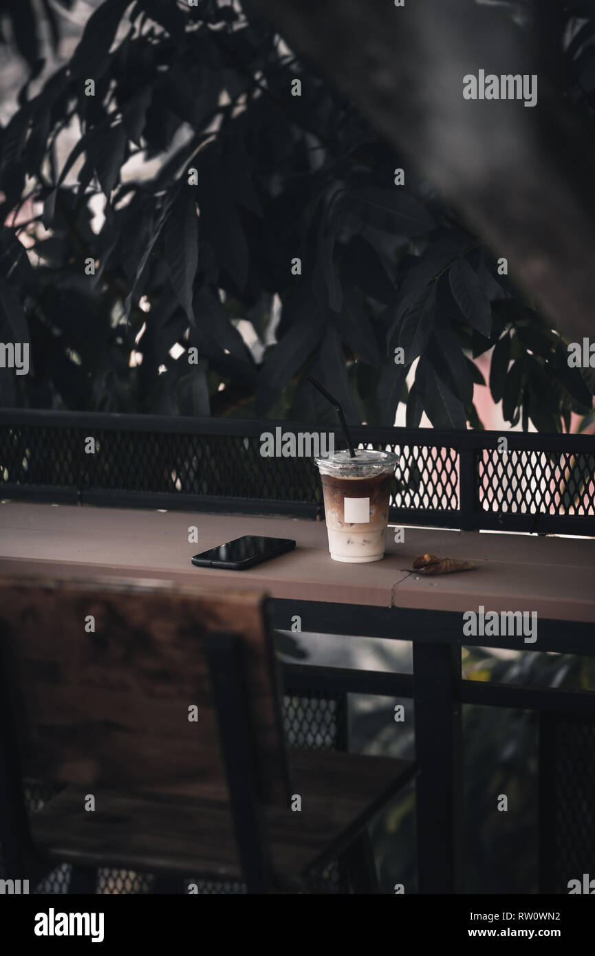 Abstrakte Szene von Iced Latte in Kunststoff Kaffeetasse mit blank white label und auf Holz bar am Nachmittag Zeit smartphone. Wochenende Aktivität und relaxatio Stockfoto