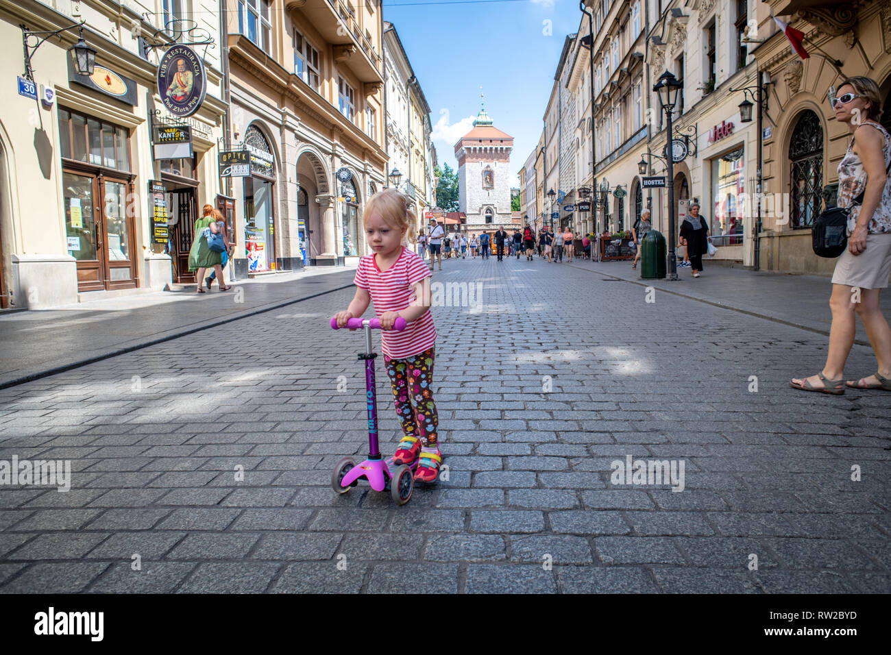Kleines Mädchen reitet auf Scooter entlang der gepflasterten Straßen der Marktplatz in Krakau - w Altstadt mit Brama Florianska, Stadtmauer Tor, hinter Ihnen Stockfoto