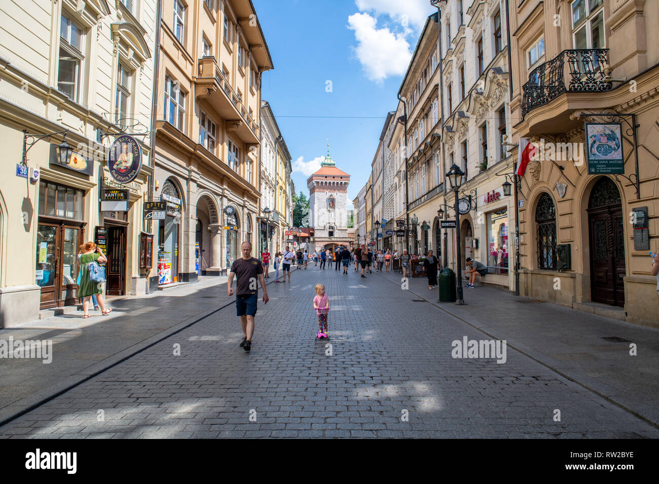Kleines Mädchen reitet auf Scooter entlang der gepflasterten Straßen der Marktplatz in Krakau - w Altstadt mit Brama Florianska, Stadtmauer Tor, hinter Ihnen Stockfoto