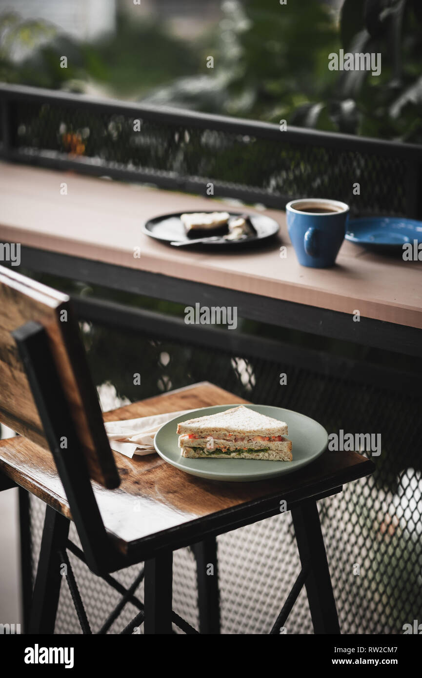Abstrakte Szene von Frühstück Mahlzeit einschließlich Thunfisch Sandwich, einen langen schwarzen Kaffee und Schokoladenkuchen Käsekuchen auf Holz im Außenbereich bar. Wochenende Morgen Acti Stockfoto