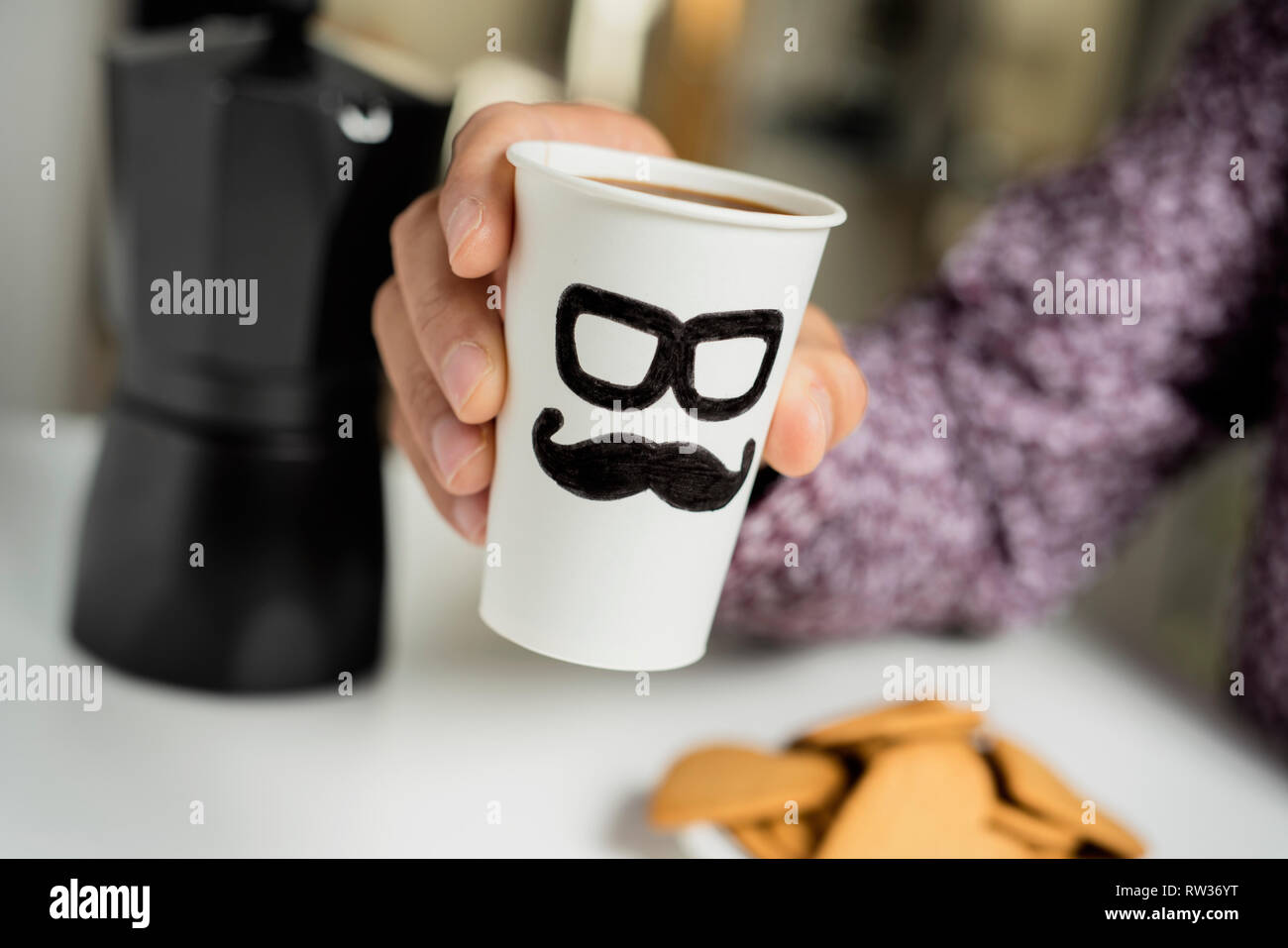 Nahaufnahme von einem kaukasischen Mann mit einer Wegwerf- Tasse mit Kaffee in seinen Händen, mit ein paar Gläser und einen Schnurrbart in es gezeichnet, an einem Tisch sitzen f Stockfoto