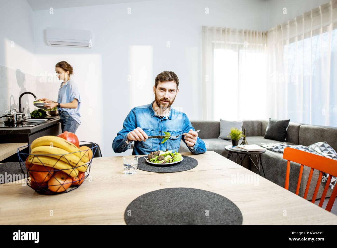 Man gesund essen Salat im Wohnzimmer der modernes Apartment mit Frau Geschirr auf dem Hintergrund Stockfoto