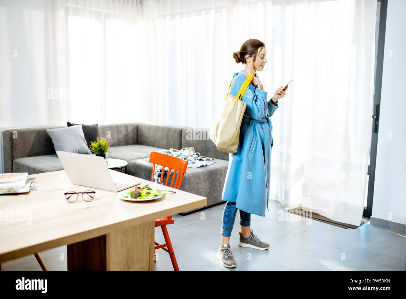 Junge stilvolle Frau im Mantel mit Telefon und Tasche im modernen Apartment Bereit zum Verlassen Stockfoto