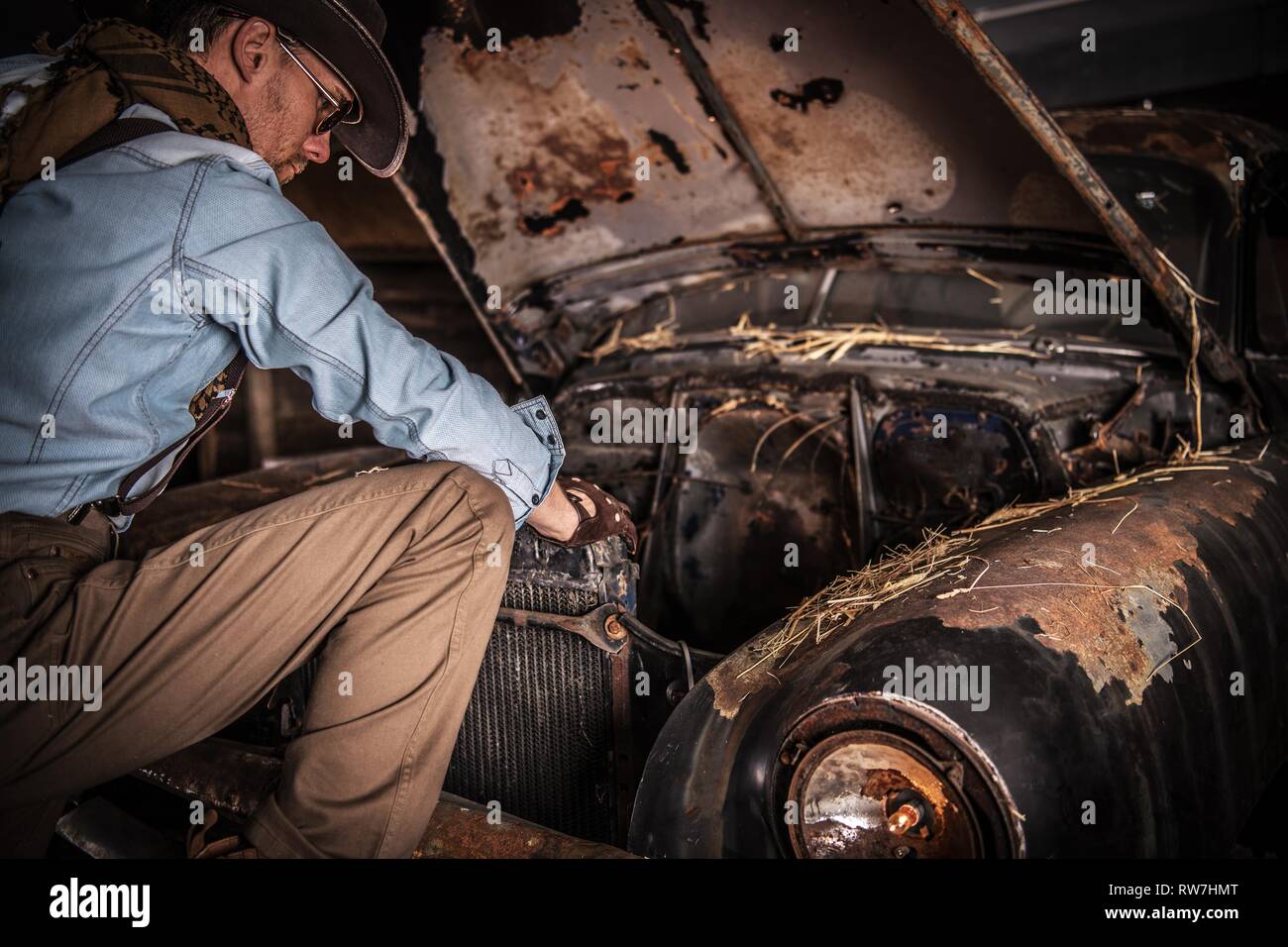 American Cowboy und den rostigen Klassiker in der alten Scheune. Kaukasische Männer tragen im westlichen Stil hat sich unter der Motorhaube. Stockfoto