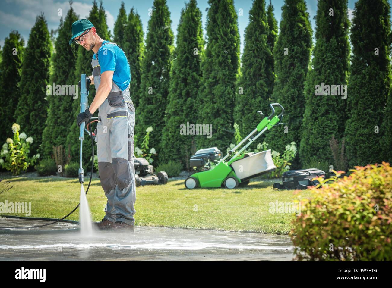 Kaukasischen Männern in seinem 30s Reinigung Gartenwege und Auffahrt mit professioneller Hochdruckreiniger. Gartengeräte im Hintergrund. Stockfoto