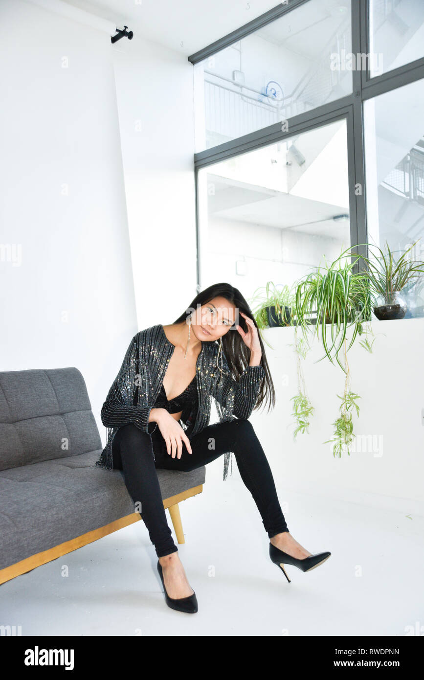 Eine schöne asiatische Mädchen mit einem sexy schwarzen Outfit ist auf einem Sofa in einem eleganten weißen Zimmer sitzen, sie ist glücklich und entspannt Stockfoto