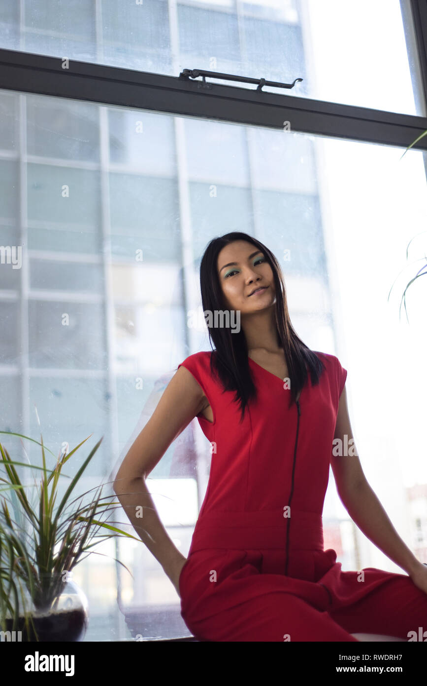 Eine schöne asiatische Mädchen im roten Overall sitzt mit dem Rücken zu einem Fenster aus Glas, von üppigen Pflanzen umgeben - Es ist ein Tower Block außerhalb Stockfoto