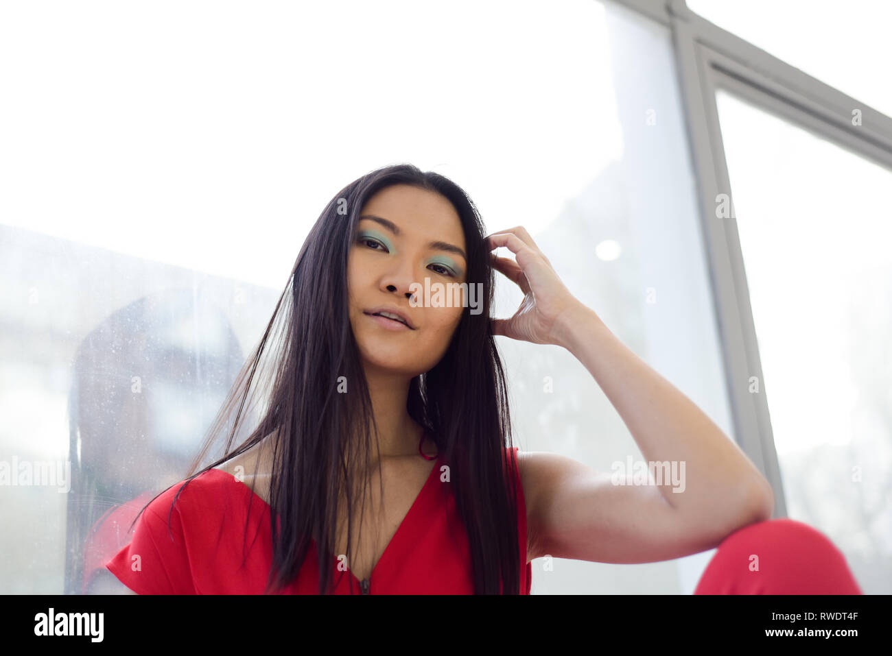 Eine schöne asiatische Mädchen im roten Overall sitzt mit dem Rücken zu einem Glas Fenster - Sie sieht nachdenklich, sie die Gebäude und die Straße sehen können Stockfoto