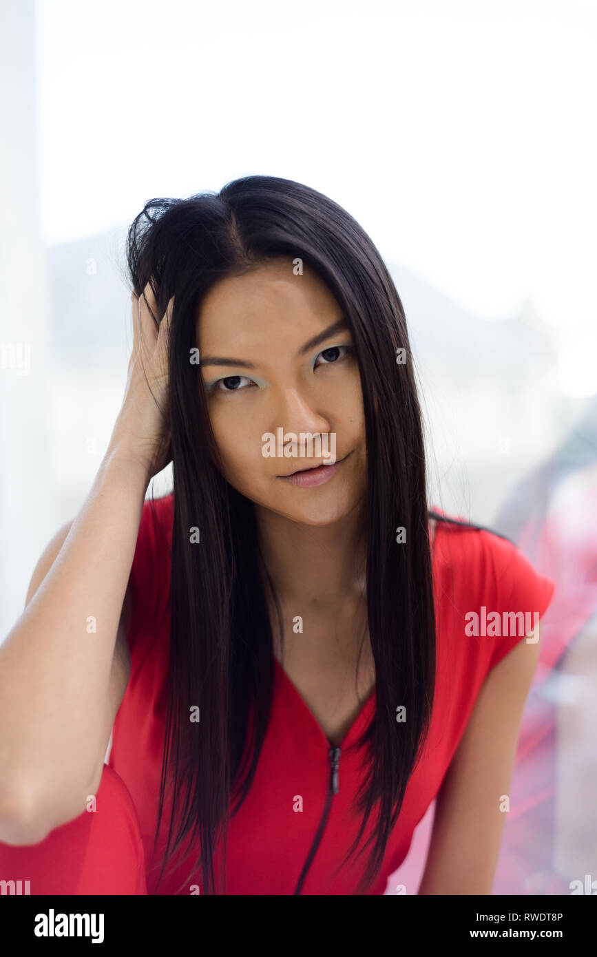 Eine schöne asiatische Mädchen im roten Overall sitzt mit dem Rücken zu einem Glas Fenster - Sie sieht nachdenklich, sie die Gebäude und die Straße sehen können Stockfoto