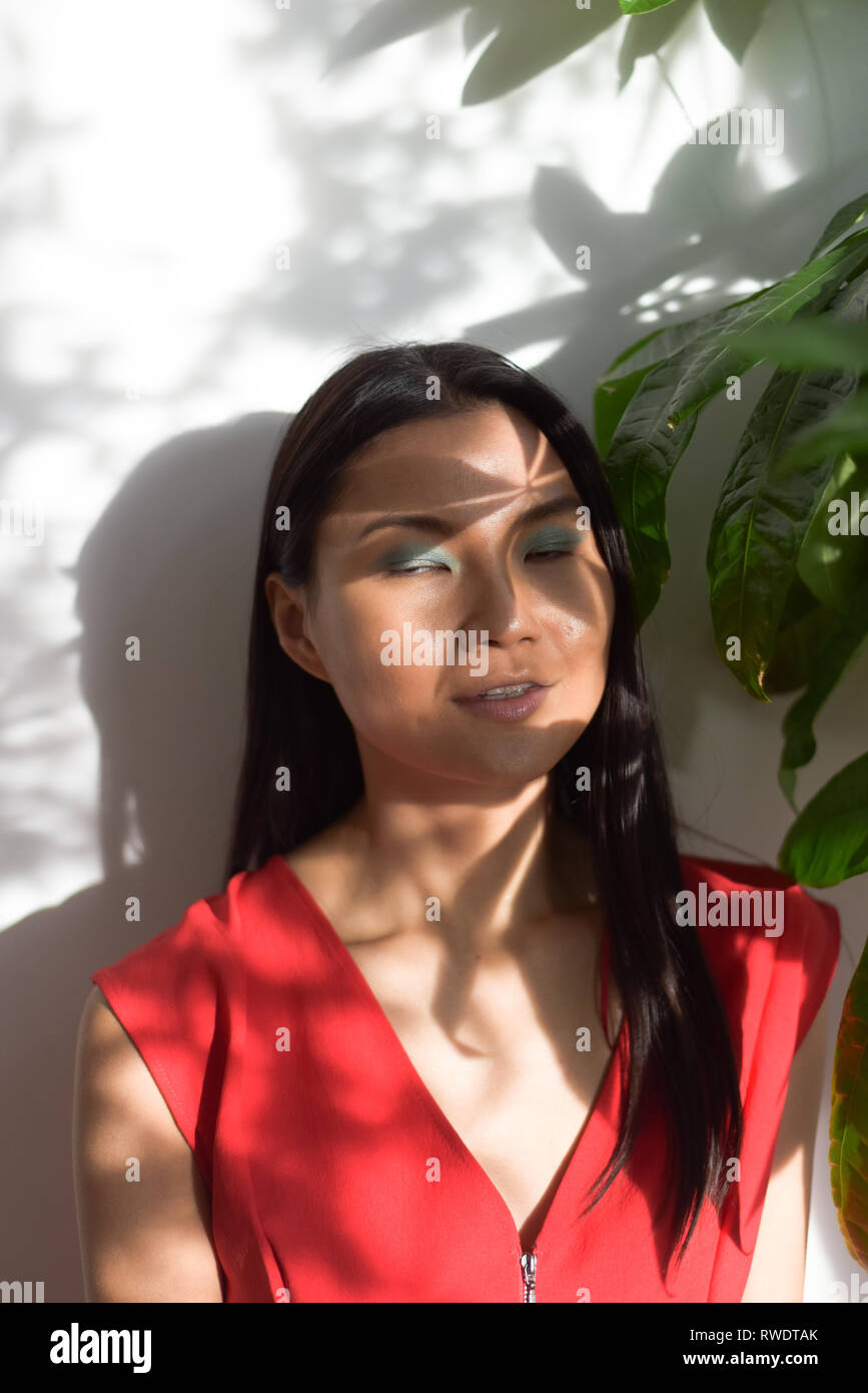 Eine schöne asiatische Mädchen mit einem roten Overall sitzt in der Nähe von einem großen, grünen Anlage und die Schatten Abdeckung ihr Gesicht Stockfoto