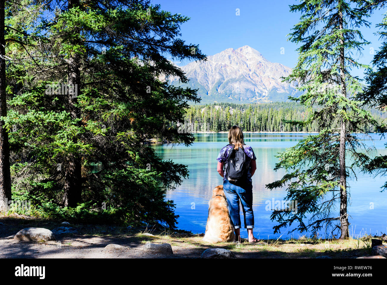 Eine Frau und ihr Golden Retriever machen eine Pause beim Wandern um den Lac Beauvert im Jasper National Park in Jasper, Alberta. Pyramid Mountain ist im Hintergrund. Stockfoto