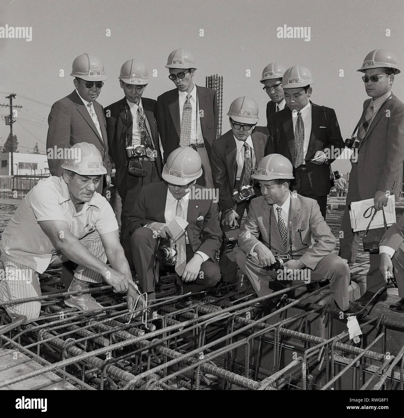1970, Los Angeles, USA, eine Gruppe von geeignet japanische Bauträger mit Schutzhelmen auf vor Ort durch ein Amerika Mitarbeiter des Robert E. McKee Bauunternehmen, die Stahlkonstruktion oder den Support für ein neues Gebäude, Stockfoto
