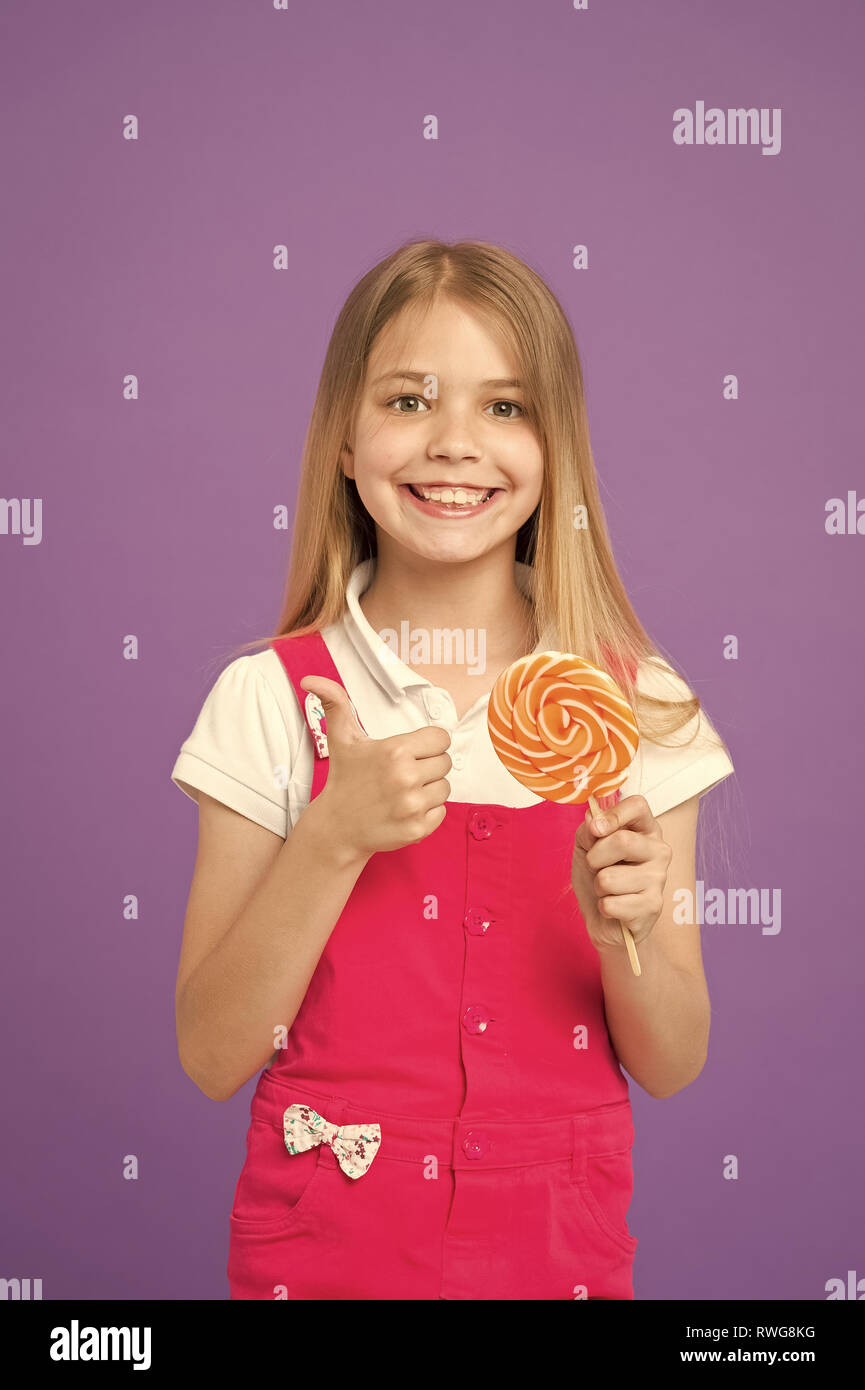 Mädchen mit bunten Lutscher. Süße Kindheit Konzept. Kind mit langen Haaren gerne Süßigkeiten. Mädchen mit lächelnden Gesicht hält Giant candy in der Hand, violett unterlegt. Kind in rosa Overalls, Daumen hoch. Stockfoto