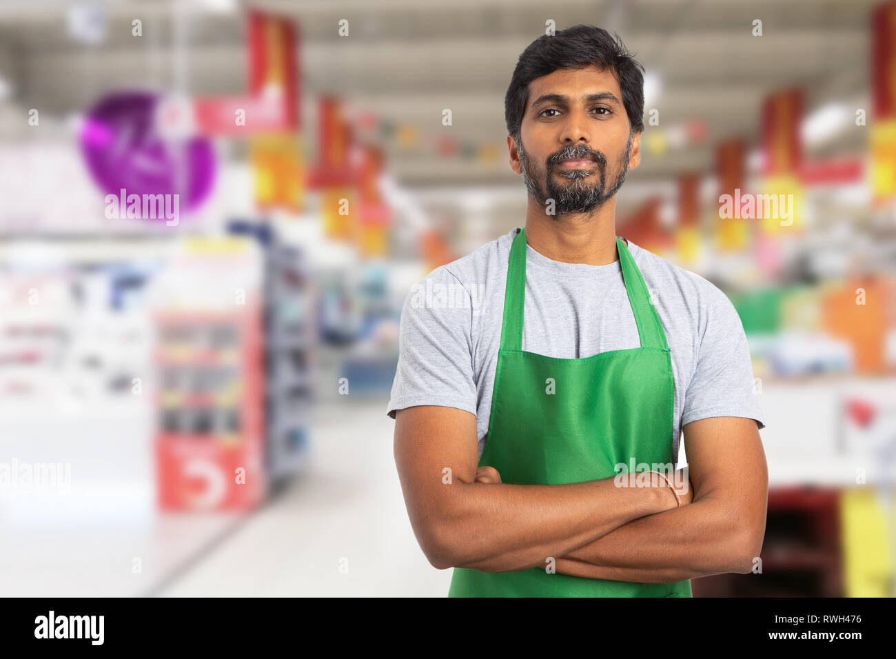 Indische männlichen Supermarkt oder SB-Warenhaus Mitarbeiter portrait Bild tragen Grüne Schürze mit verschränkten Armen und ernsten Ausdruck Stockfoto