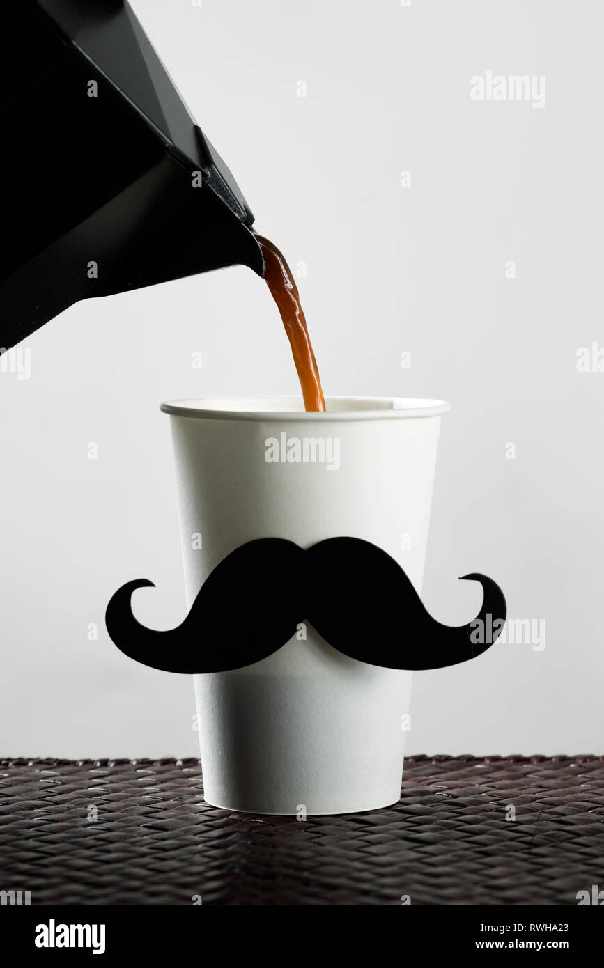 Nahaufnahme von einem weißen Einweg Becher, mit einem gefälschten Schnurrbart, gefüllt mit Kaffee aus einem schwarzen Kaffeekanne, vor einem weißen Hintergrund Stockfoto